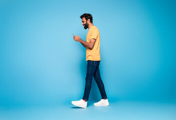Indian guy in casual outfit walking with smartphone and smiling on blue studio background, full...