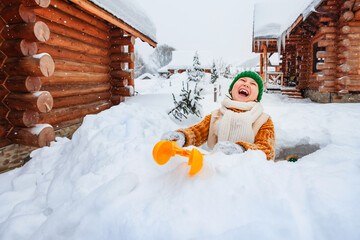 Happy smiling child playing with snow in the yard. Kid makes snowballs with hand tool.