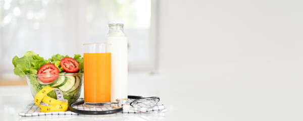 diet, healthy eating, food and weigh loss concept - close up of salad fresh  fruit ,orange juice  and measuring tape with stethoscope on white table in kitchen banner copy space