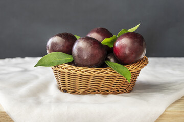 A basket full of ripe red plums with green leaves on a white cloth