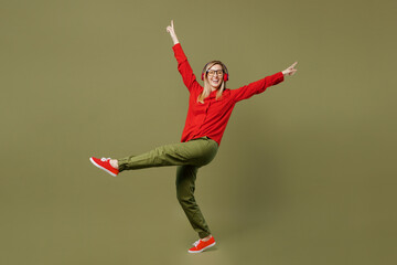 Full body side view young woman she wear red shirt casual clothes glasses listen to music in headphones rais eup legs dance isolated on plain pastel green background studio portrait Lifestyle concept