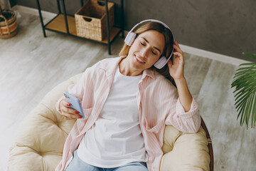 Top view young woman wearing casual clothes sits in archair listen to music in headphones use mobile cell phone stay at home hotel flat rest spend free spare time in living room indoor Lounge concept