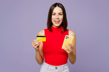 Young woman wears red tank shirt casual clothes using mobile cell phone hold credit bank card doing online shopping order delivery booking tour isolated on plain pastel light purple background studio.