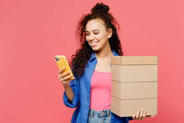 Young woman of African American ethnicity she wear blue shirt casual clothes hold stack cardboard blank boxes use mobile cell phone isolated on plain pastel pink background studio. Lifestyle concept.