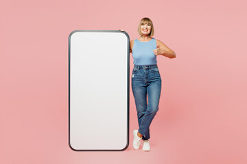 Full body elderly fun blonde woman 50s years old she wears blue undershirt casual clothes big huge blank screen mobile cell phone smartphone with area show thumb up isolated on plain pink background.