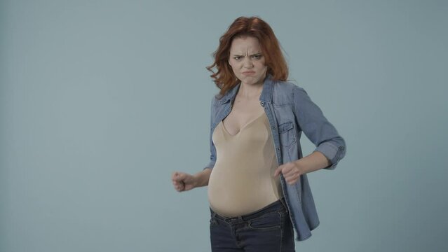 A redhaired pregnant woman strokes her bloated belly with a smile, gets angry, upset, offended. Woman in the studio on a blue background. Pregnancy and emotions, changeable mood, hormones.