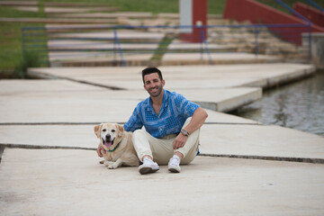 Handsome young man with beard and his Labrador retriever dog, posing for photos by a lake in the...