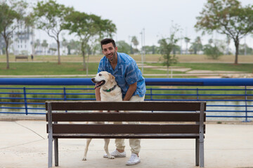 Fototapeta premium Handsome young man with beard and his Labrador retriever dog on a wooden bench in the park. The man and his pet are looking happily at the camera. Pets and pets concept