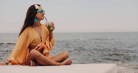 Beautiful young woman in boho styled crop top drinking cocktail and smiling while relaxing seaside