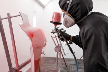Car body shop technician in full coveralls adjusting a pressurized paint spray gun in a painting booth