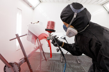 A male worker in jumpsuit and blue gloves paints with a spray gun a side part of the car body in red after being damaged at an accident. Auto service industry professions