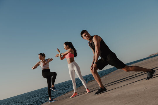 Three young people in sportswear doing stretching exercises before training seaside together