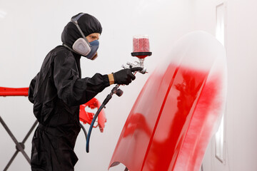 Car painter in protective clothes and mask painting a car, mechanic using a paint spray gun in a...