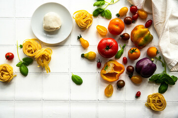 Ingredients for vegetable pasta.
Dry tagliatelle nests, variety of fresh tomatoes, peppers, basil and mozzarella ball on white tile background.