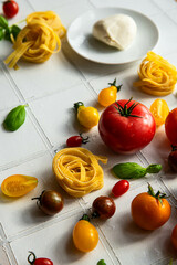 Ingredients for vegetable pasta.
Dry tagliatelle nests, variety of fresh tomatoes, peppers, basil and mozzarella ball on white tile background.