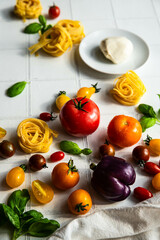 Ingredients for vegetable pasta.
Dry tagliatelle nests, variety of fresh tomatoes, peppers, basil and mozzarella ball on white tile background.