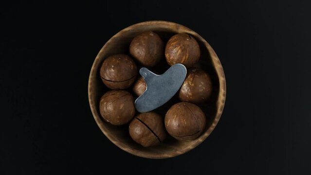Macadamia nuts with sawn nutshell and special metal key used to dry open the nut. Wooden bowl with Australian Macadamia on dark background, top view. 4K video, Rotating.