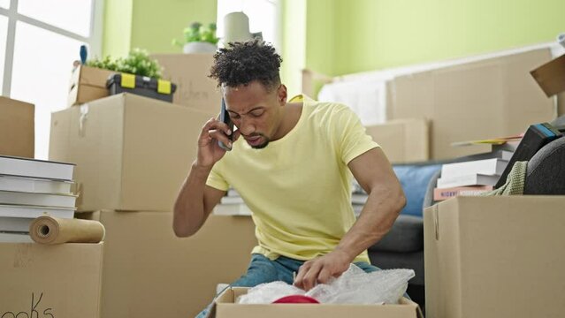 African american man unpacking cardboard box talking on smartphone looking upset at new home