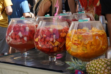 Market stand with pineapple and punch drinks. Row of serving bowls full of punch with various types of fruits. Beside there is a stack of plastic glasses. Defocused incidental people are walking by. 