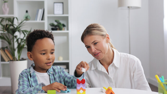 African American child curly little boy and child development specialist blonde woman playing with colorful wooden bricks sitting at table, making pyramid together. Exercises for children with autism.