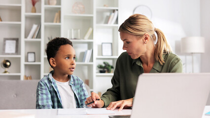 Blonde tutor teaching little African American preschooler to write at home. Help with early childhood education in an online school. Happy boy is learning to write.