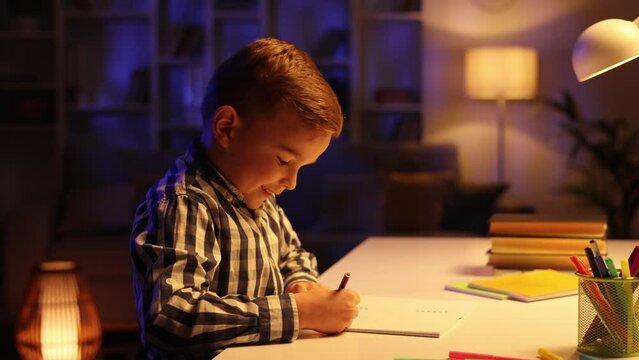 A little brunette boy sits at the table in his room in the evening and draws. A preschooler uses his imagination, creates a picture, dreams of being an artist.