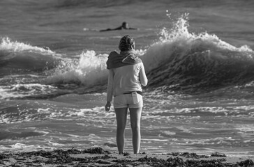Fototapeta premium A woman on a Vendée beach watches surfers on the ocean, dressed in shorts with a smartphone in the back pocket, a small towel on her back, black and white photo.