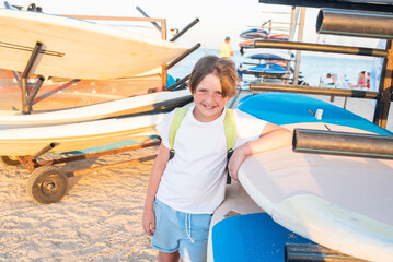 Fototapeta na wymiar smiling child in a t-shirt with a backpack at the surf station on the beach in summer. joyful 10 year old child stands next to the windsurf boards, waiting for a workout.