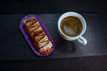 Cup of coffee and delicious eclair cake - tiramisu flavoured covered with cream, icing and chopped nuts, on a black background.