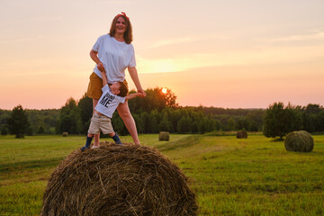 Naklejka na ściany i meble Happiness is found in the simplicity of nature as a woman with baby boy standing on a hay bale during sunset. Portrait of a happy kid two years old and a woman thirty five years old