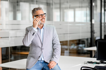 Stylish mature businessman in smart casual wear talking on the smartphone in the office in relaxed atmosphere, male manager, ceo, employee leaned on the desk and having pleasant phone conversation