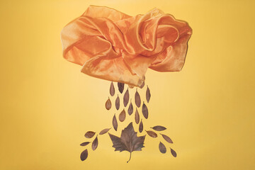 Minimal autumn season concept with a rainy weather storm made with an orange textile cloud and autumnal dried leaves as rain drop. composition on yellow wallpaper or background.