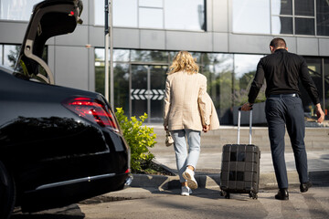 Chauffeur helps a businesswoman to carry her suitcase into a building, walking together, luxury...