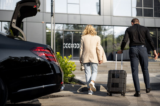 Chauffeur helps a businesswoman to carry her suitcase into a building, walking together, luxury taxi in front. Concept of business trips, idea of a luxury car transfer service