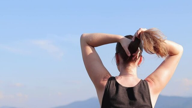 Girl tourist straightens her hair, hot sunny day. Girl against the blue sky. A woman in nature breathes fresh air, rear view. Hair with a crease from an elastic band