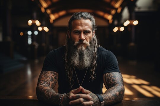 Portrait of an old man with a long gray beard and mustache in a shirt with tattoos on his arms sitting on the floor in a dark room