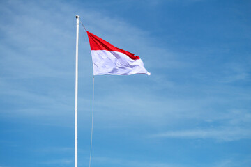 The Indonesian flag flutters on a bamboo pole against a blue sky background, waving the red and white flag