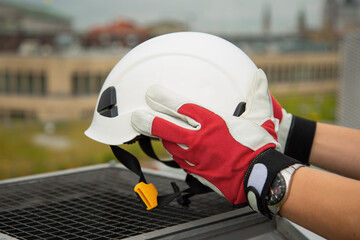 Hands of a worker in protective gloves hold a protective helmet over a ventilation grill on a city background