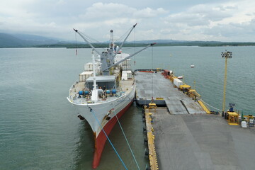 Naklejka premium White refrigerated cargo ship moored in port of Puerto Barrios. Reefer vessel is older style bulk vessel with derricks. Behind is coastline of Guatemala.