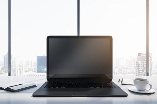 Close up of laptop with empty mock up monitor, coffee cup and notepad placed on desk. Window with bright city view in the background. 3D Rendering.