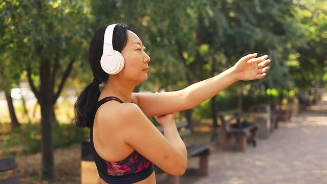 Middle age asian woman wearing sportswear and headphones stretching arms at park
