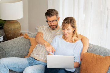 Middle-aged couple wearing casual clothes and sitting at home on the sofa and using a laptop - 639975406