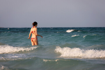 Slim girl in bikini going to swim in blue sea waves. Beach vacation in stormy weather