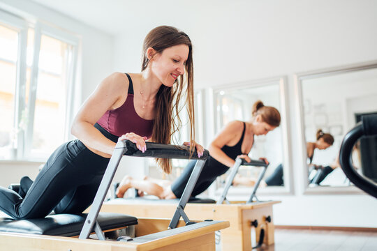 Two young females doing ABS muscles strength exercises using  pilates reformer machine in sport athletic gym hall. Active people training, yoga classes concept.