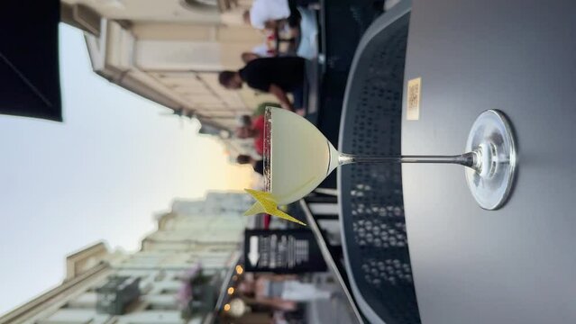 A classic alcoholic gin based cocktail Corpse Reviver in a coupe glass on a table of a terrace of a cafe or bar in old town. People, tourists in bokeh walking by. Lifestyle vertical video.