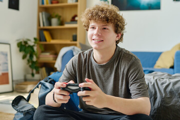 Cute smiling teenage boy with joystick pressing buttons during computer game while sitting by bed in front of camera and enjoying leisure