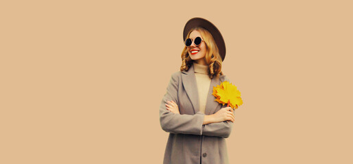 Autumn color style outfit, portrait of beautiful smiling young woman with yellow maple leaves wearing round hat, coat on brown background