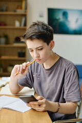 Brunette teenage girl in grey t-shirt watching online video in smartphone while sitting by desk in front of camera and thinking of new ideas