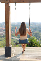 A beautiful girl is sitting on a swing. The girl rides on a swing against the backdrop of the city, an observation deck. Cityscape from the hill