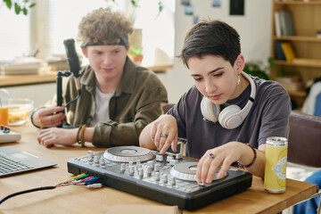 Cute teenage girl with headphones on neck adjusting turntables while sitting by desk next to her boyfriend preparing microphone for stream